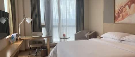 A hotel room with a bed, a desk, a chair, a small table, and a window with curtains.