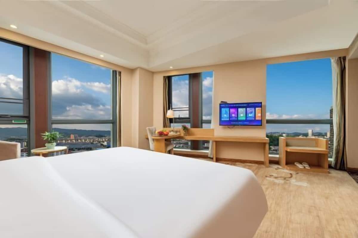 A hotel room with a large bed, a desk, a chair, a TV, and large windows with a city view.