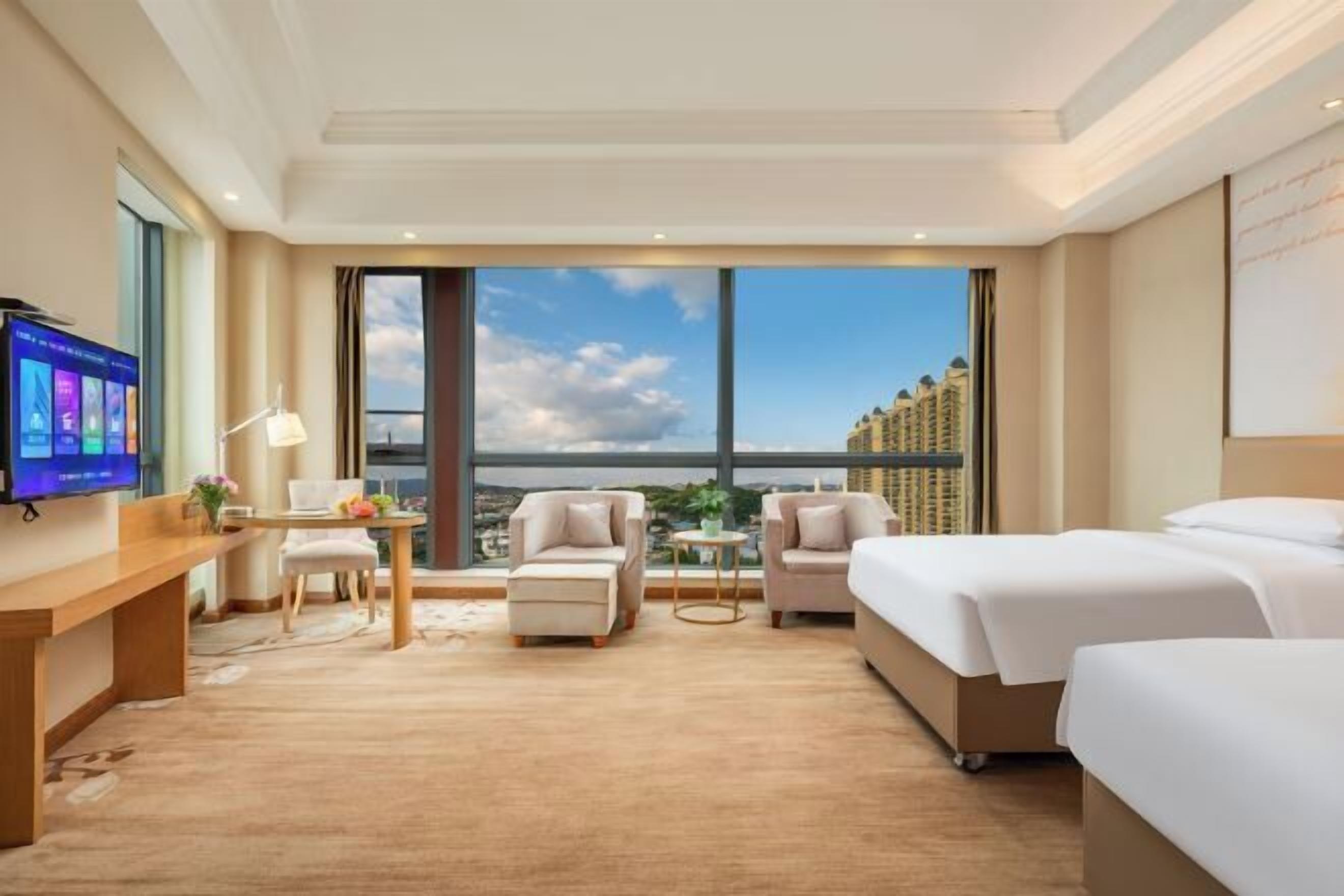 Una habitación de hotel con un ventanal grande, un escritorio con computadora, dos sillones y dos camas.