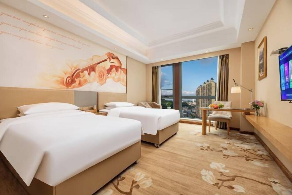 A hotel room with two beds, a desk, a chair, a TV, and a large window with a city view.