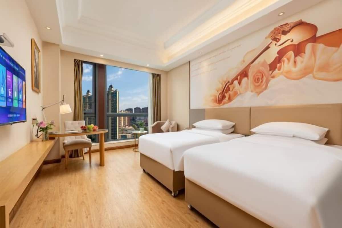 A hotel room with two beds, a desk with a chair, a TV, and a large window with a city view.