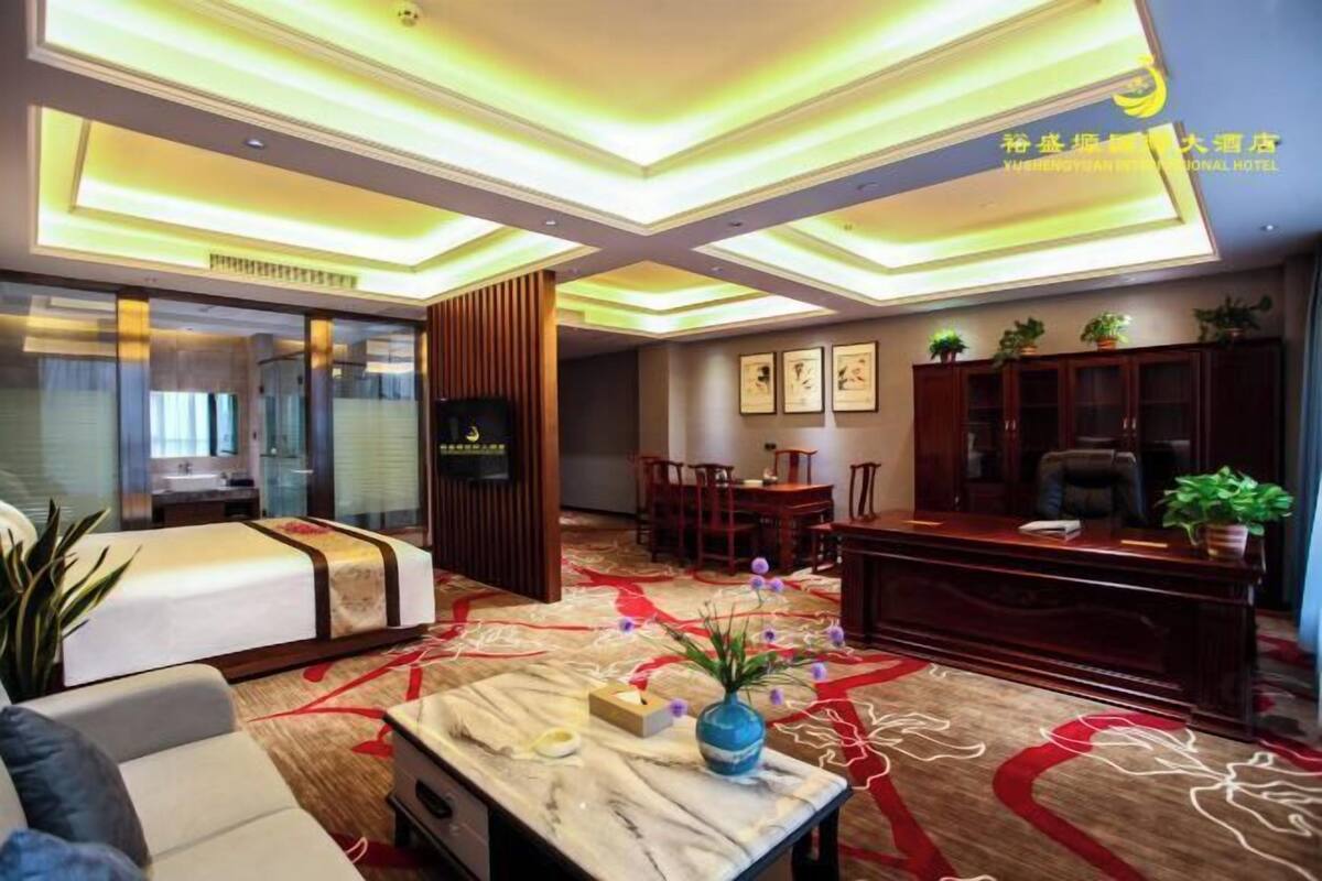 Habitación de hotel con cama, escritorio, silla, mesa con un florero y alfombra de diseño rojo y blanco.