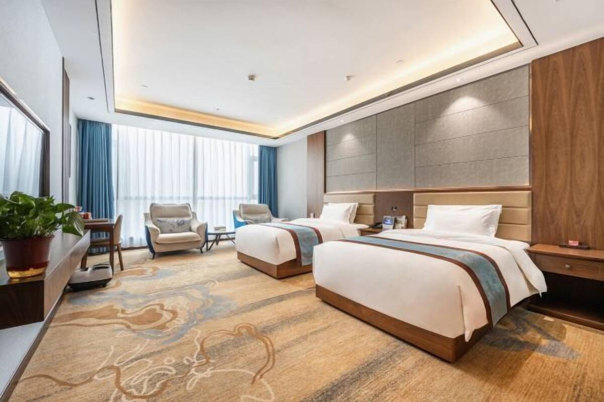 Habitación de hotel con dos camas, un escritorio con silla, un ventanal con cortinas y un cabecero moderno.