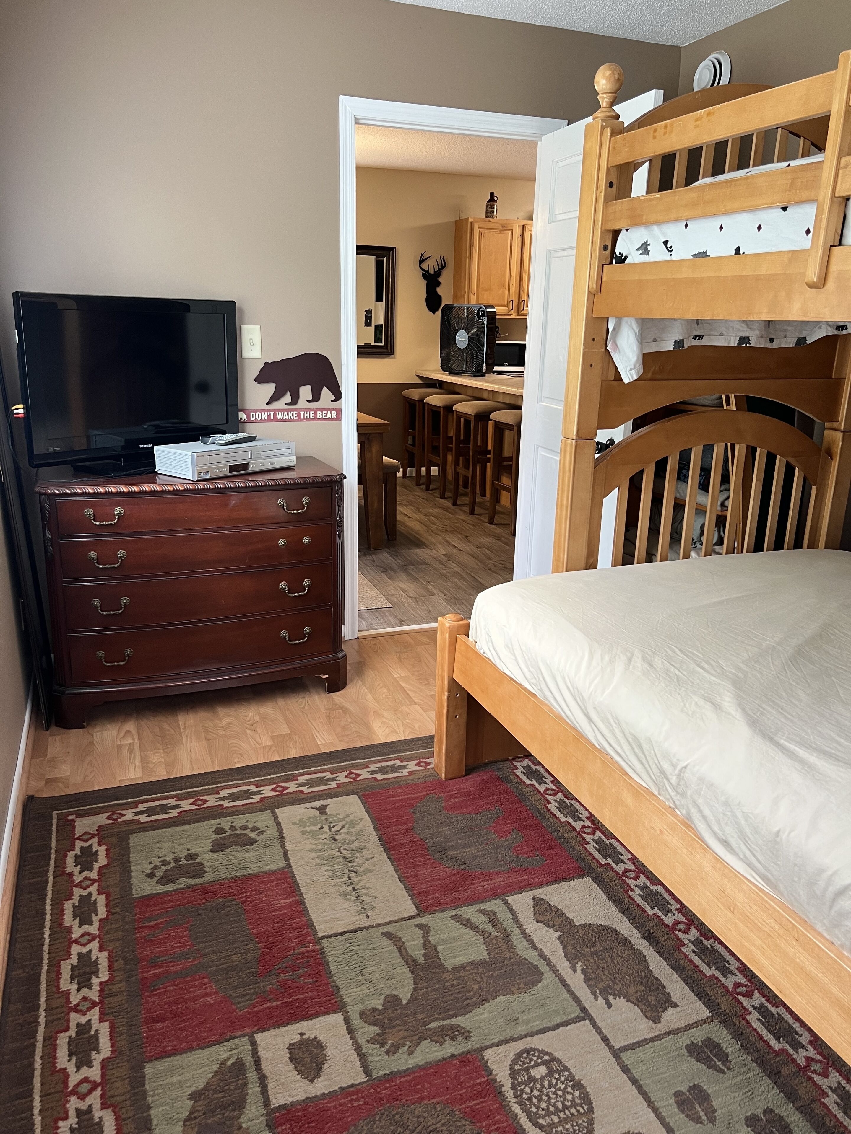 Un dormitorio con una cama nido de madera, televisión, cómoda y una cocina al fondo.