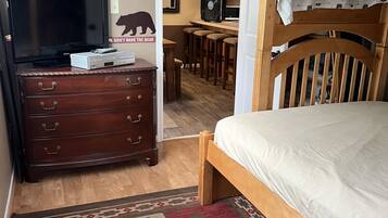 Un dormitorio con una cama nido de madera, televisión, cómoda y una cocina al fondo.