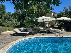 A swimming pool with lounge chairs and umbrellas, surrounded by greenery and a building in the background. - Holiday apartment with a view and pool (Jadranovo)