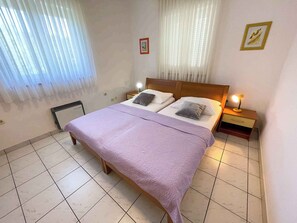 A bedroom with a bed, two bedside tables, a heater, and a window with sheer curtains.