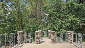 Un pont en pierre, doté de garde-corps métalliques, qui mène à une zone boisée.