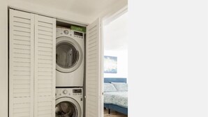 A laundry room with two stacked washing machines, a bed with a blue headboard, and a wooden floor. - WindnSealeep (La Jolla)