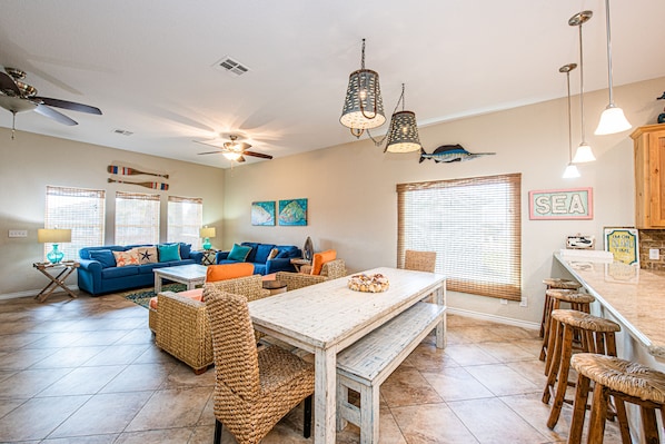 A spacious living area with a blue sofa, wicker chairs, a wooden dining table, and a kitchen area with a bar.