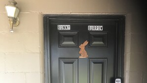 Une porte noire avec les panneaux « BUNNY » et « BURROW », une découpe en bois représentant une main, un clavier à code et un luminaire.