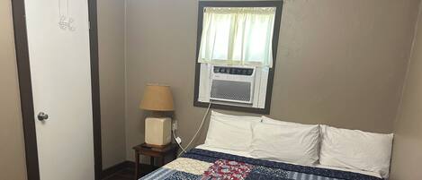 A bedroom with a bed, a nightstand, a lamp, a window with curtains, and a wall-mounted air conditioner.