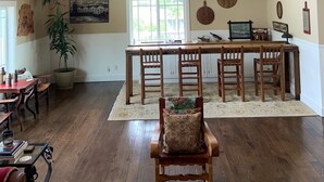 Een ruime woonkamer met een hoge plafond, houten balken, een plafondventilator, een eettafel met stoelen, een groot raam en een potplant.
