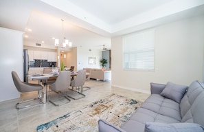 A modern living room with a grey sofa, a dining area with a table and chairs, and a kitchen in the background.