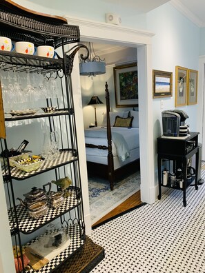 A room with a checkered floor, a bed with a wooden headboard, a coffee machine, and a shelf with various kitchen items.