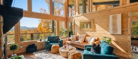 A spacious room with large windows, a cozy seating area with a sofa and chairs, a wooden ceiling, and a view of autumn trees.