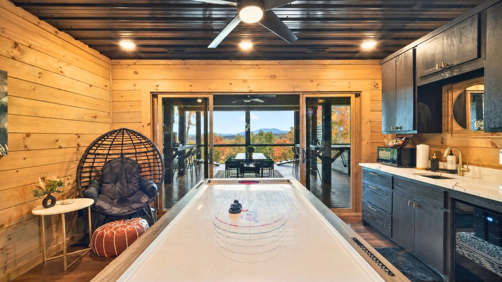 Une pièce comprenant une grande table en bois, un fauteuil confortable, une petite table sur laquelle est posé un vase, et une vue sur une terrasse extérieure, accessible par une porte vitrée.
