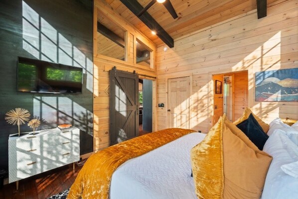 Interno di una baita in legno con un letto, una TV a parete e un quadro appeso.