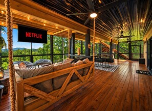 A cozy, open-plan living area with a large TV screen displaying 'NETFLIX,' wooden flooring, and a view of lush greenery.