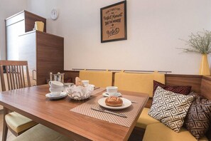 Een zithoek met een houten tafel gedekt voor twee personen, een koffiepot, kopjes en een gebakje. Op de muur hangt een ingelijste quote, en een vaas met planten staat ernaast.