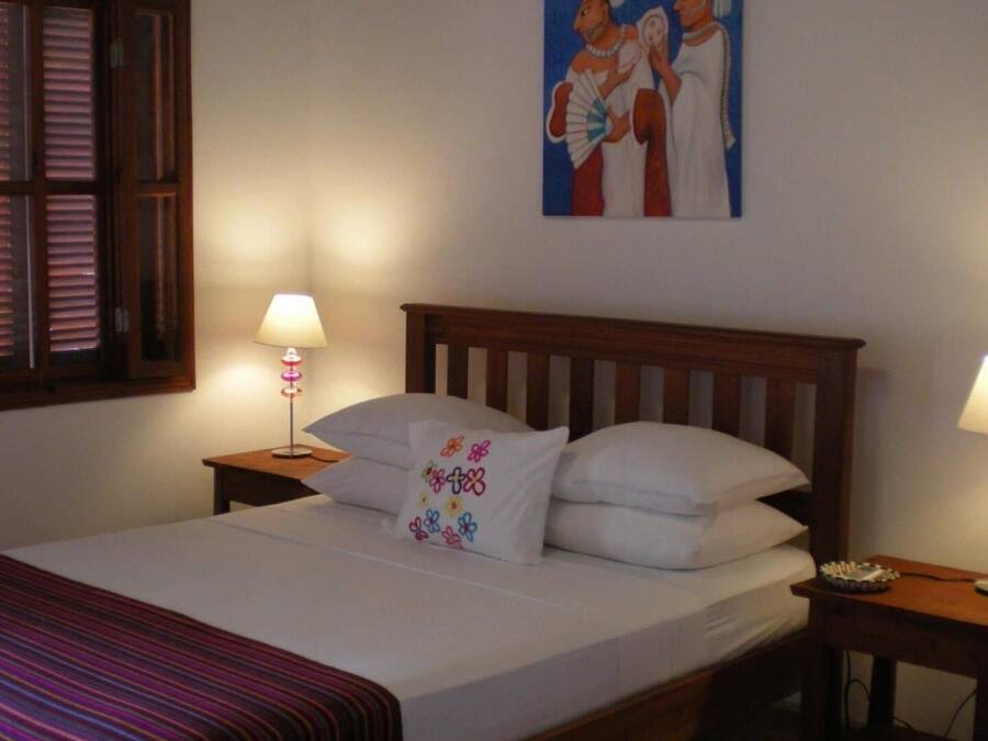 Cama bem arrumada com lençóis brancos e um travesseiro colorido, cabeceira de madeira, luminária de cabeceira e um quadro na parede.