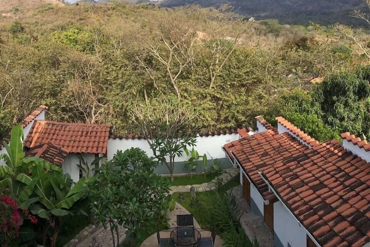 A view of a hillside with a small courtyard featuring a tiled-roof building, surrounded by greenery and mountains in the background.