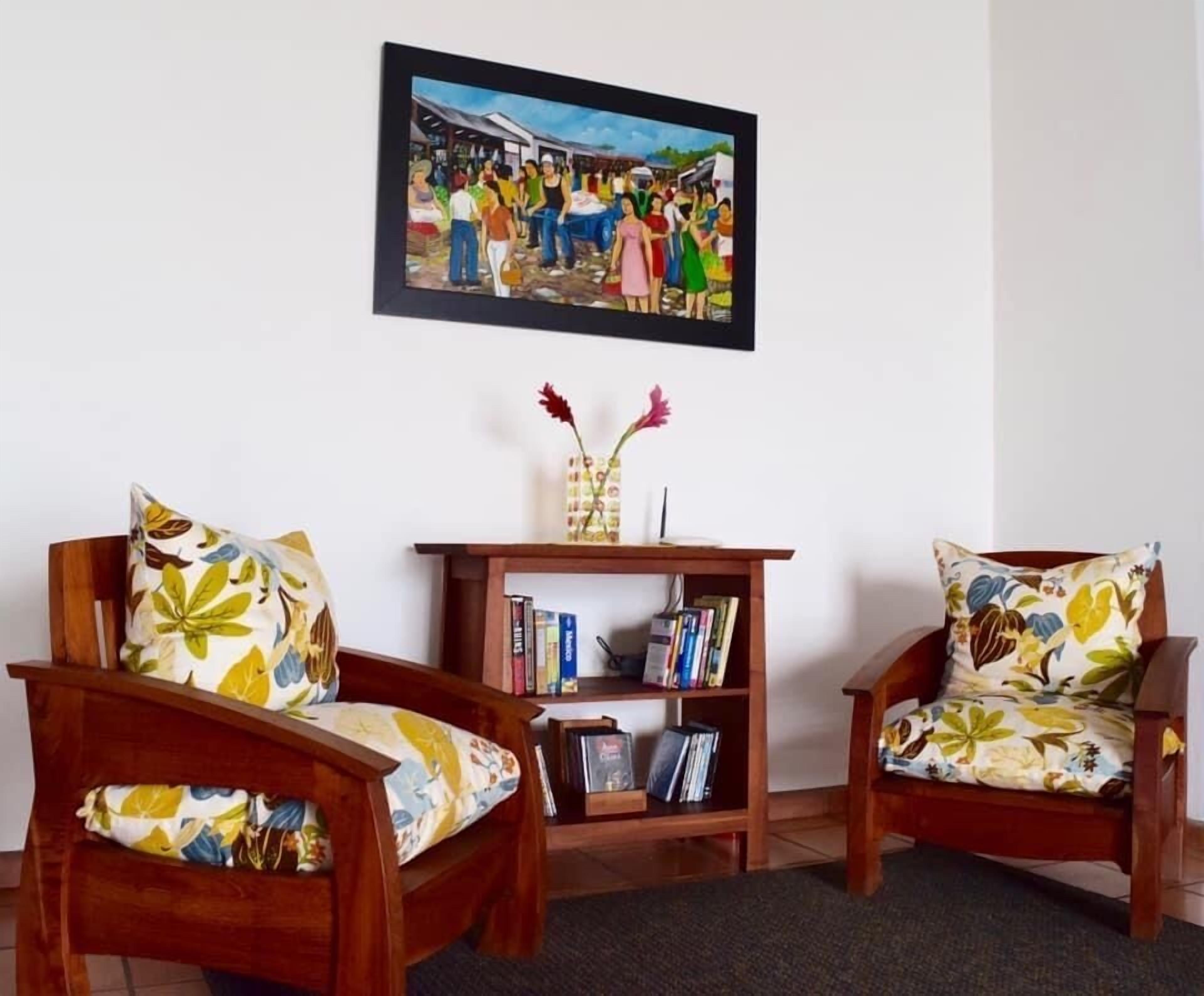 Ett vardagsrum med två trästolar med blommiga kuddar, en bokhylla och en inramad tavla på väggen.