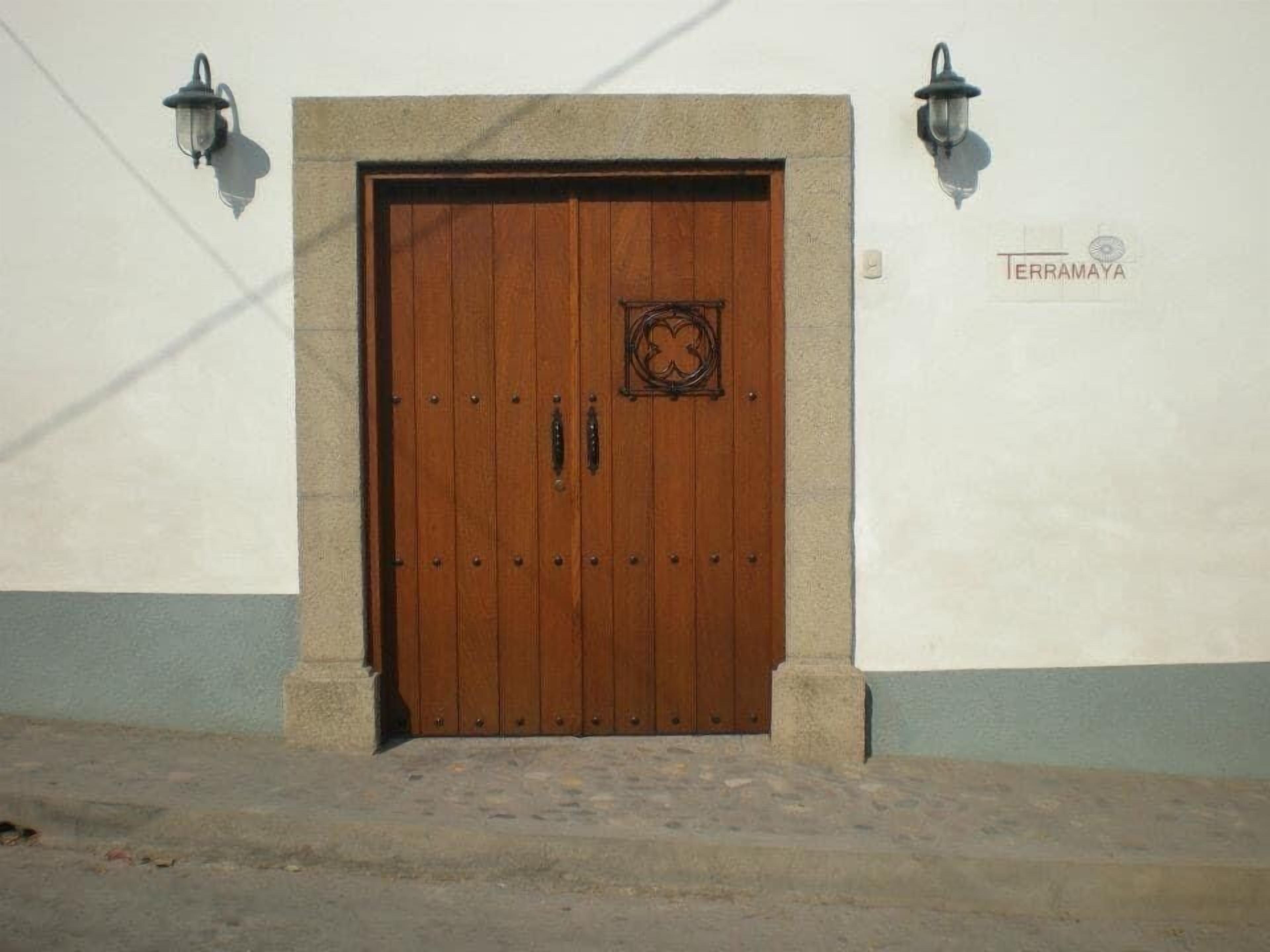 Una puerta de madera con un emblema decorativo, flanqueada por dos faroles, sobre un muro blanco con una franja azul.