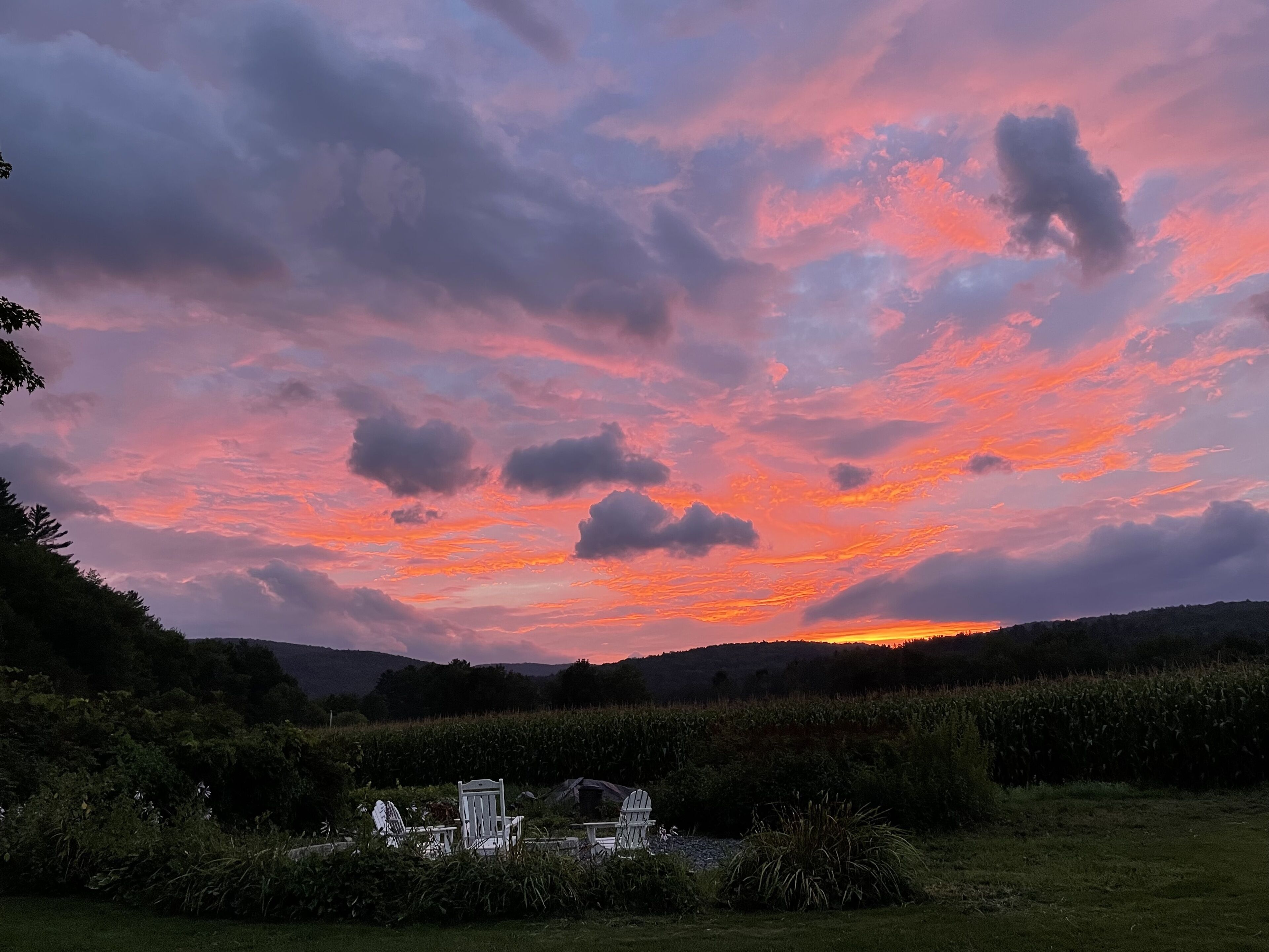 Ein Sonnenuntergang über einer ländlichen Landschaft, im Vordergrund eine Reihe weißer Stühle und ein kleiner Tisch.