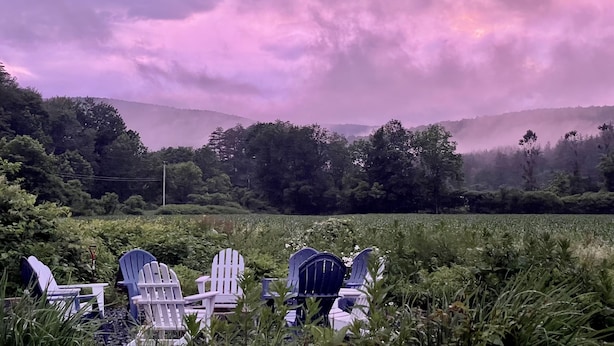 A row of Adirondack chairs in a lush green field with a backdrop of mist-covered hills and a purple sky.