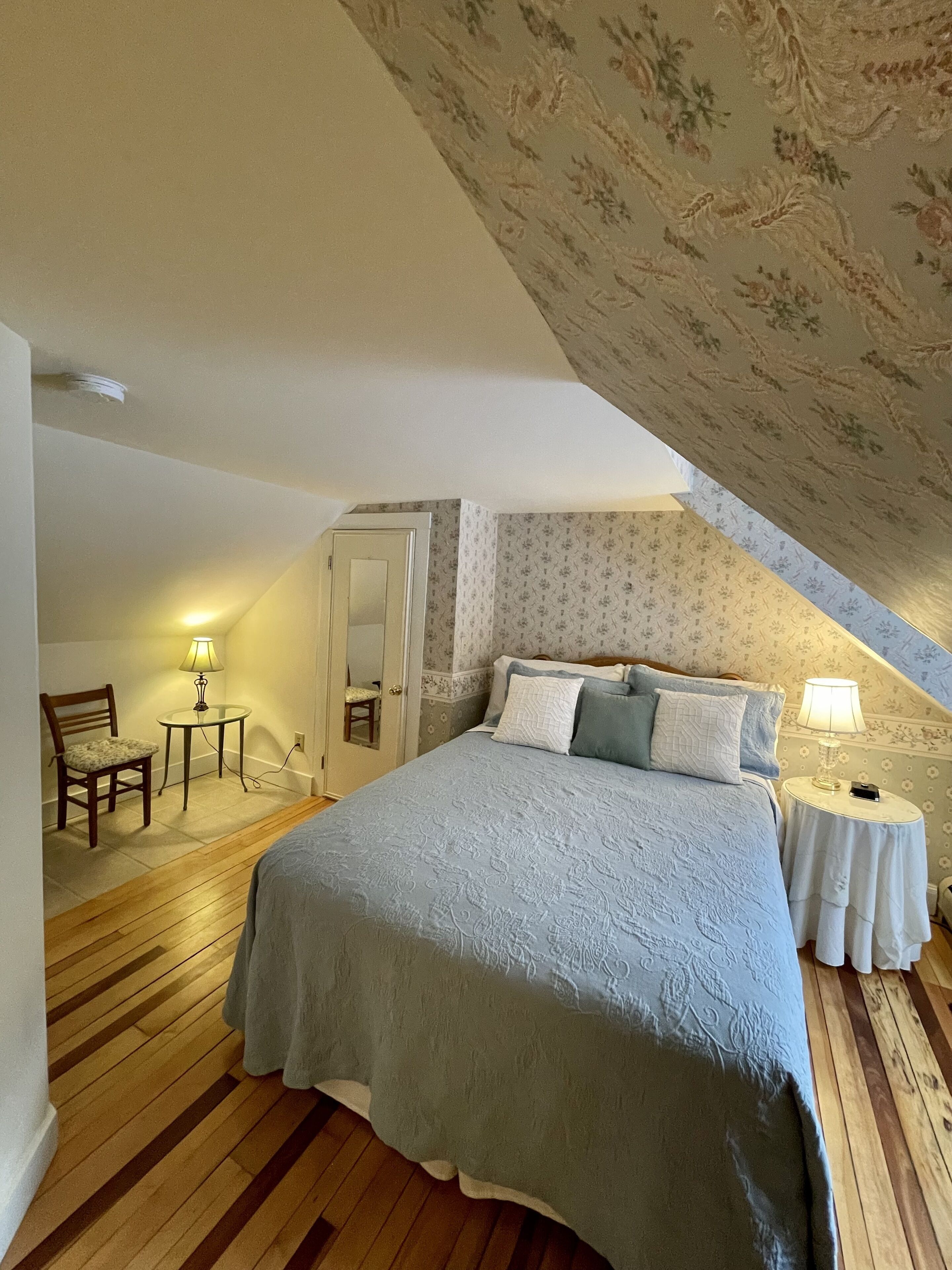 A bedroom with a sloped ceiling, a bed with a blue bedspread, two bedside tables with lamps, a chair, and a small table with a lamp.
