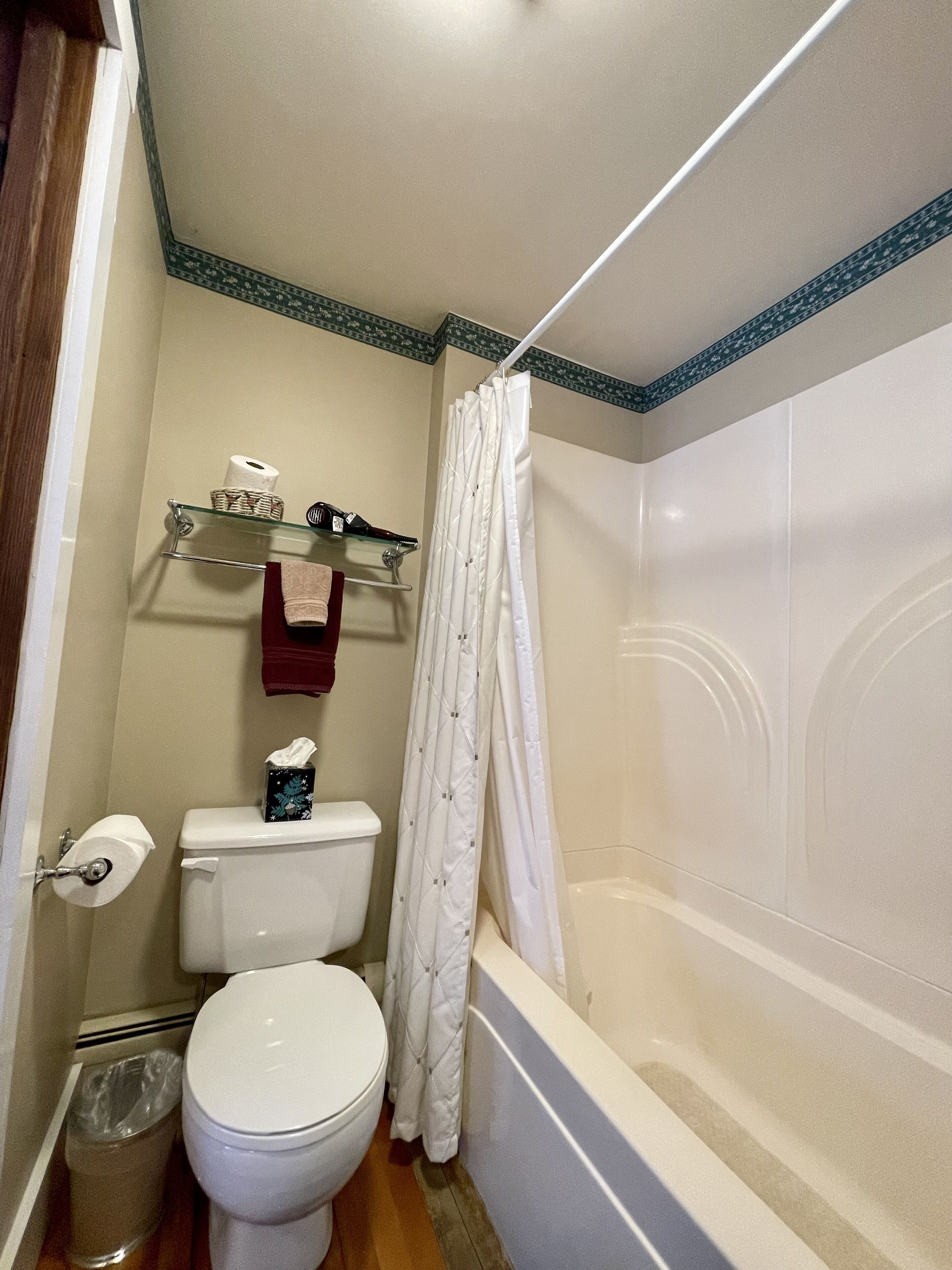 Un baño con inodoro, estante de vidrio con artículos de tocador, cortina de ducha y bañera.