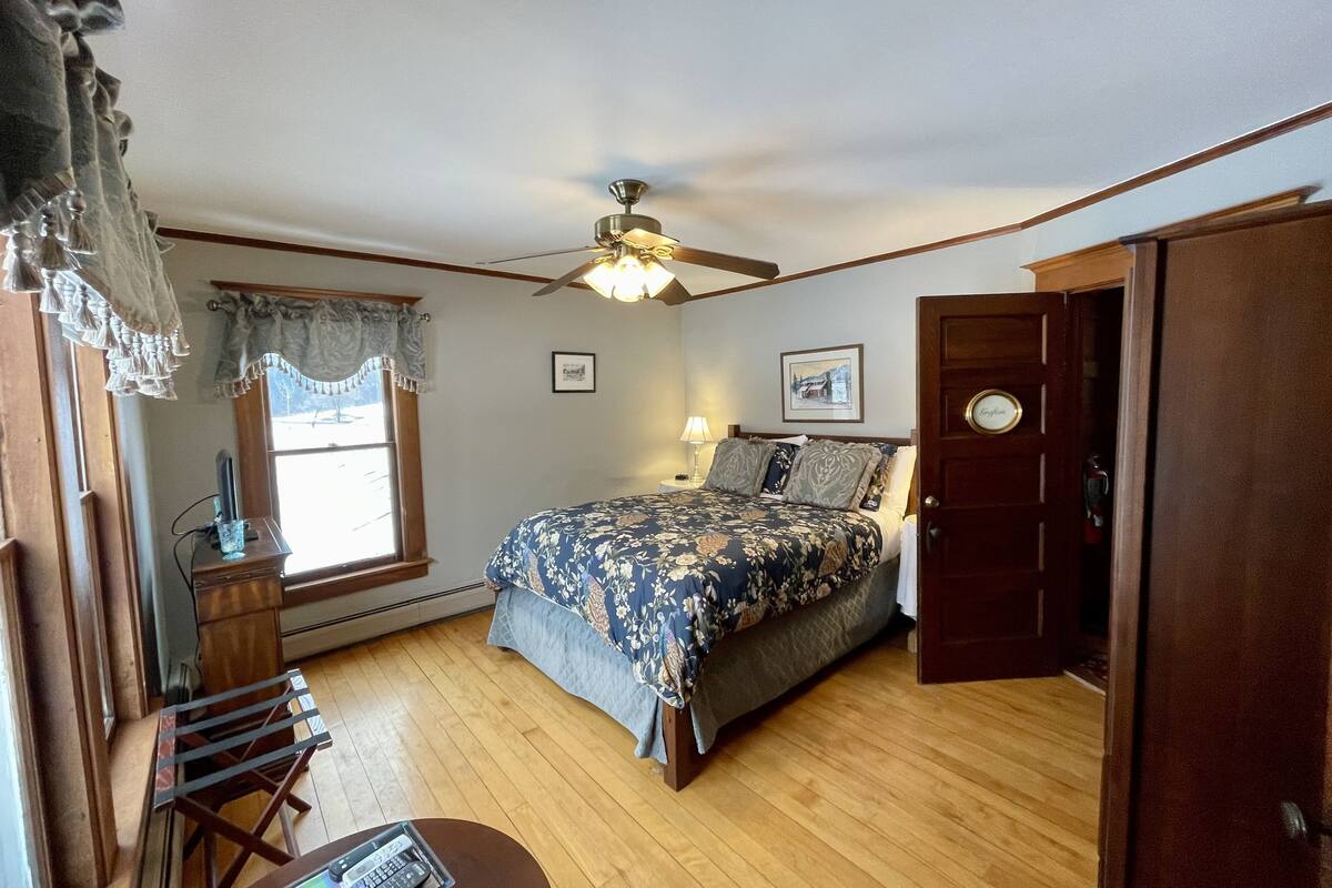 A bedroom with a bed, a ceiling fan, a window with curtains, a desk with a lamp, and a door with a nameplate.