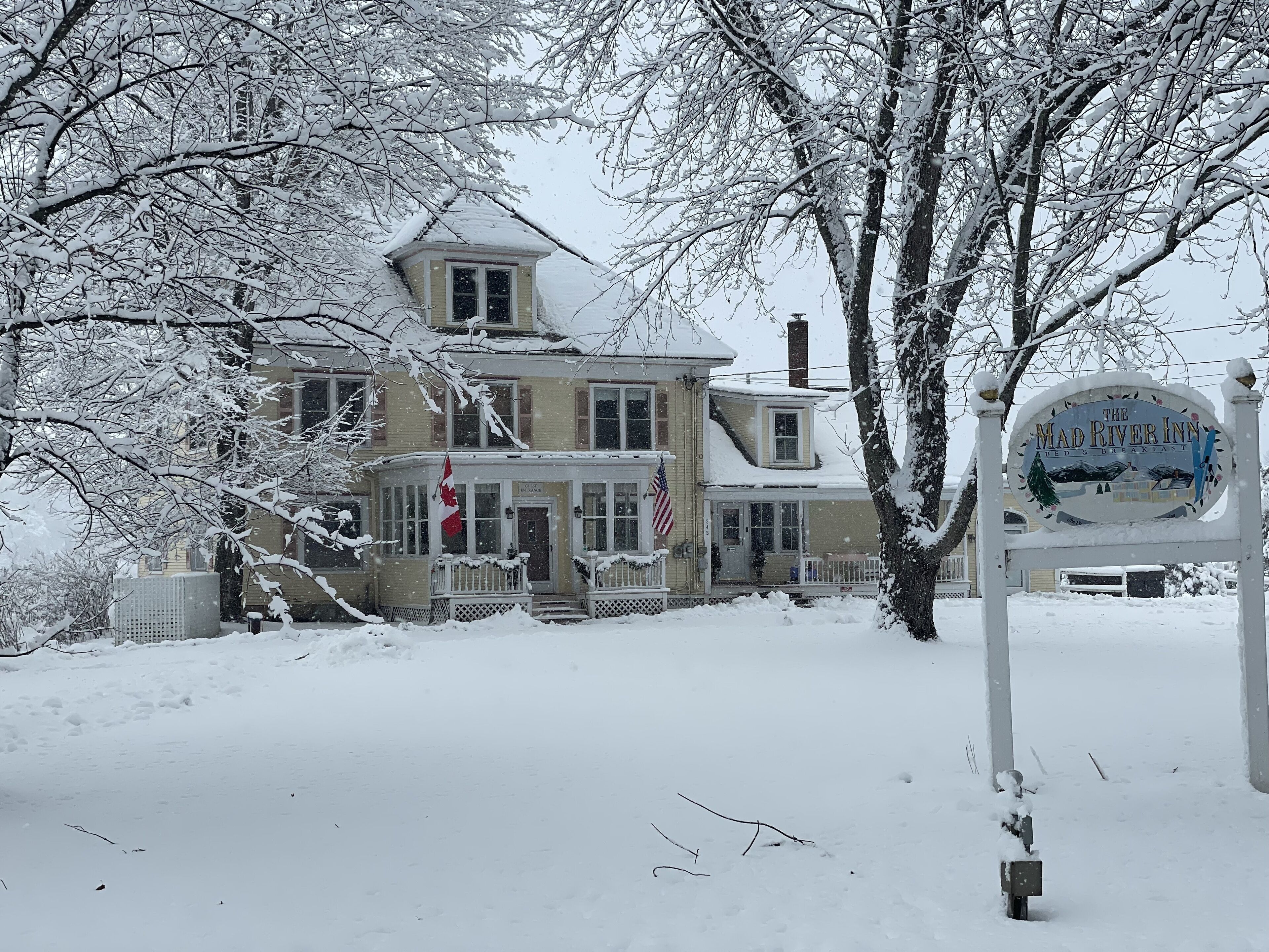 A large, two-story house with a snow-covered roof and a signboard in front.