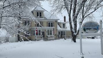 Une grande maison à deux étages, dont le toit est recouvert de neige, et devant laquelle se trouve une enseigne.