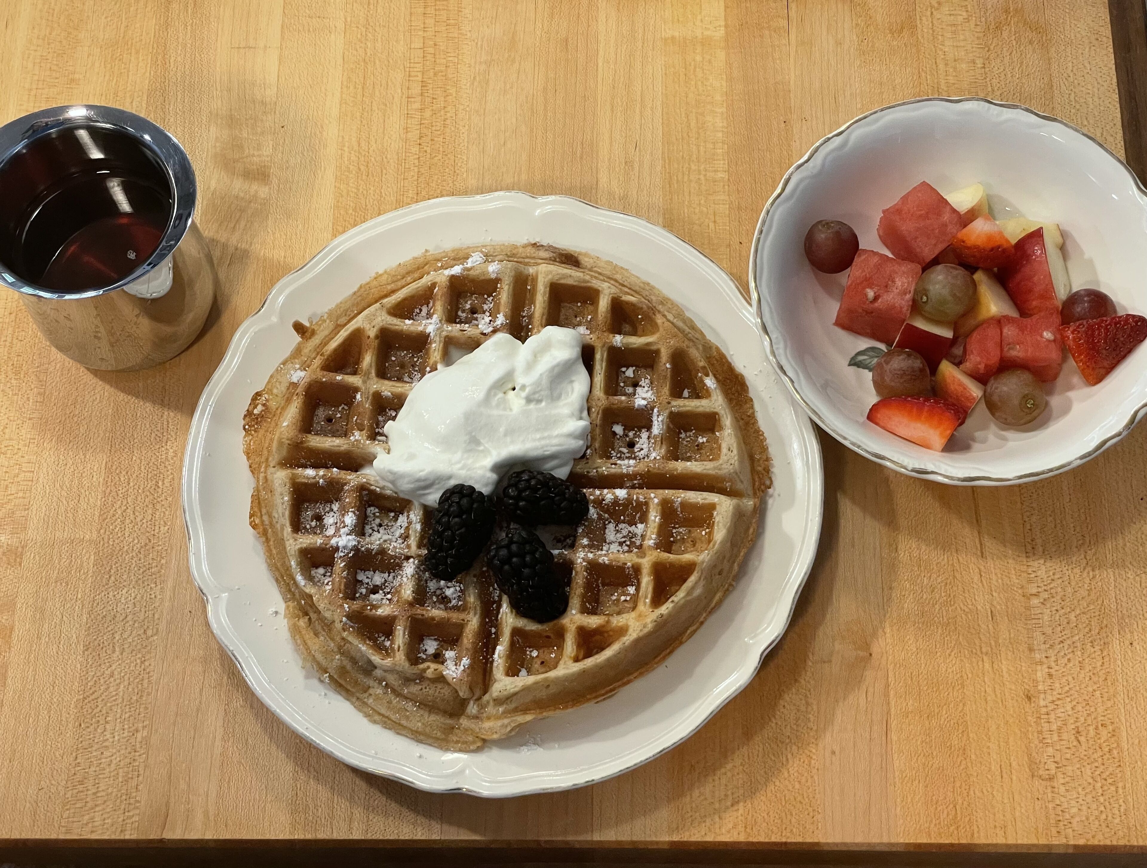Een bord met een wafel, een kom fruit en een kop koffie op een houten tafel.