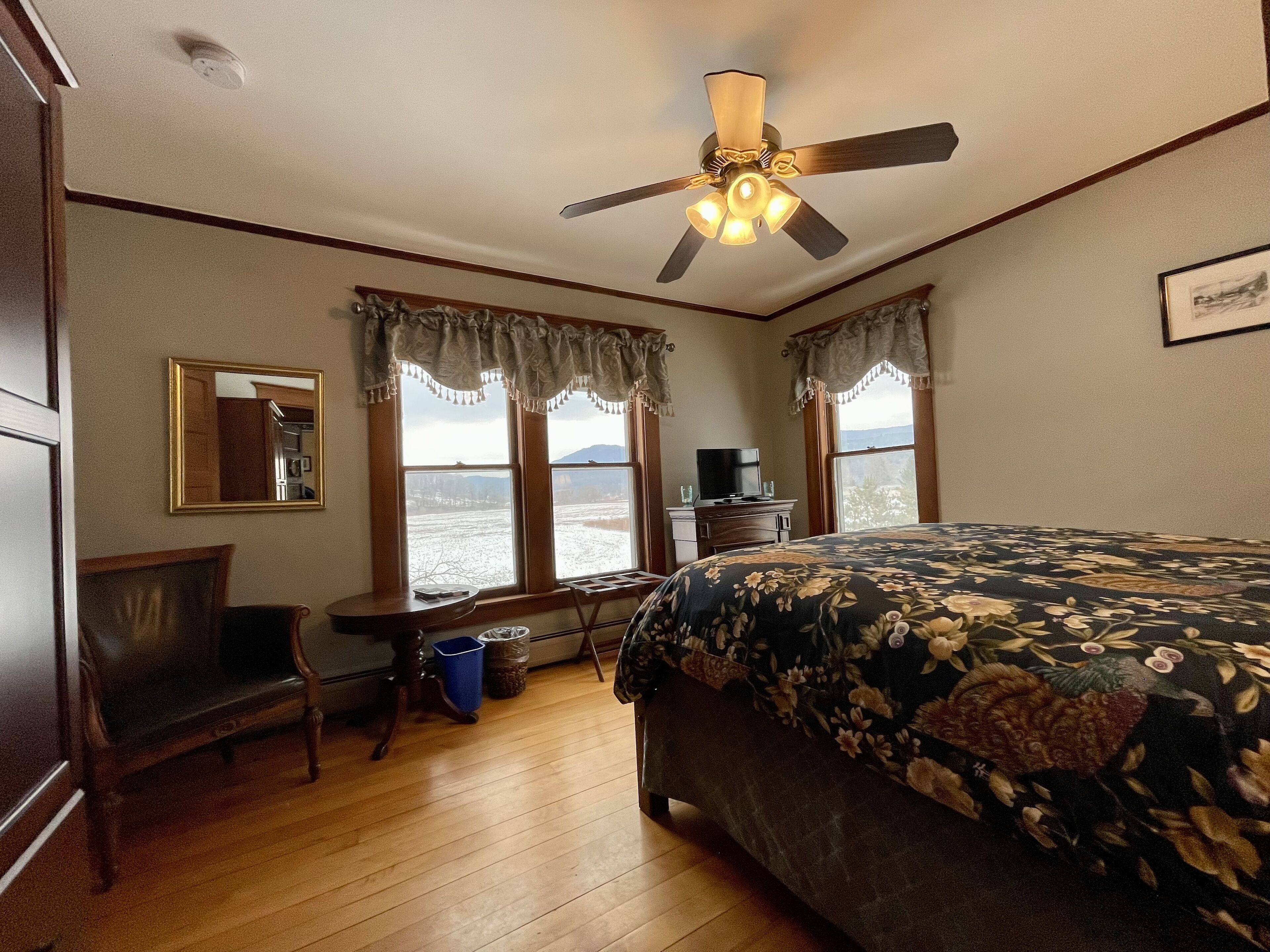 A bedroom with a bed, a desk, a chair, a ceiling fan, and large windows with curtains.