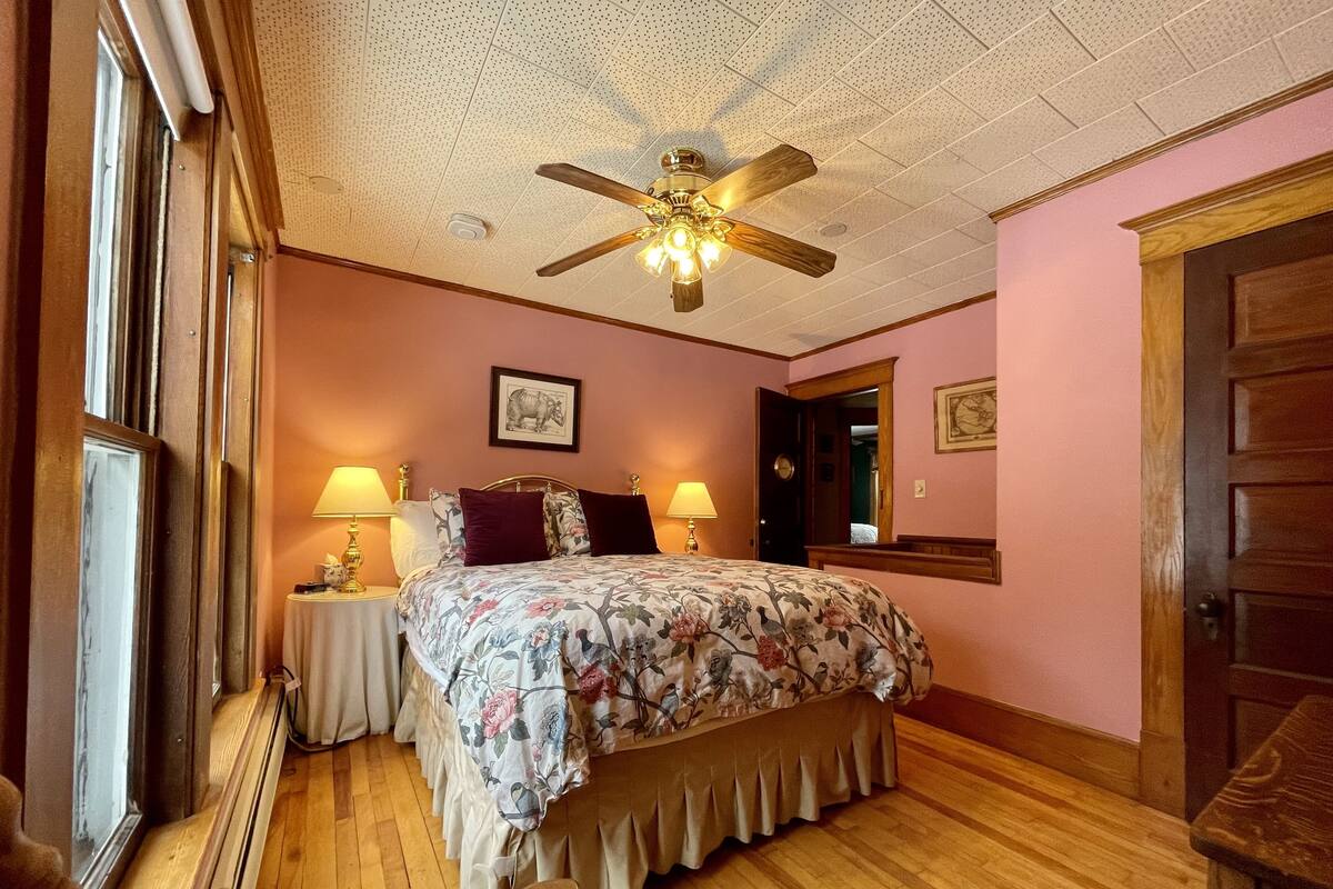 A bedroom with a wooden floor, a bed with floral bedding, two bedside lamps, a ceiling fan, and a window.