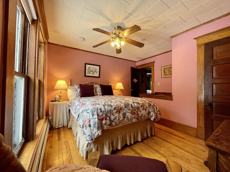 A bedroom with a wooden floor, a bed with floral bedding, two bedside lamps, a ceiling fan, and a window.