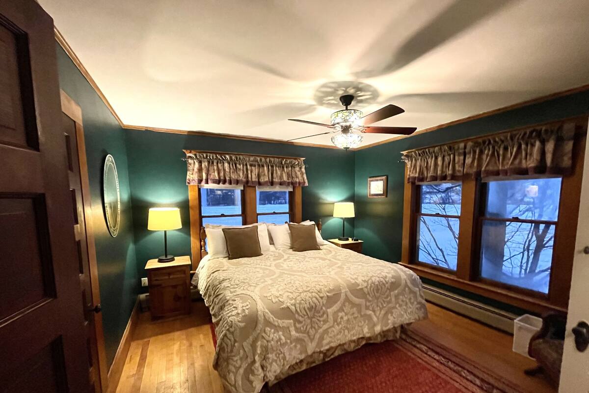A bedroom with a large bed, two bedside tables with lamps, a ceiling fan, and windows with curtains.