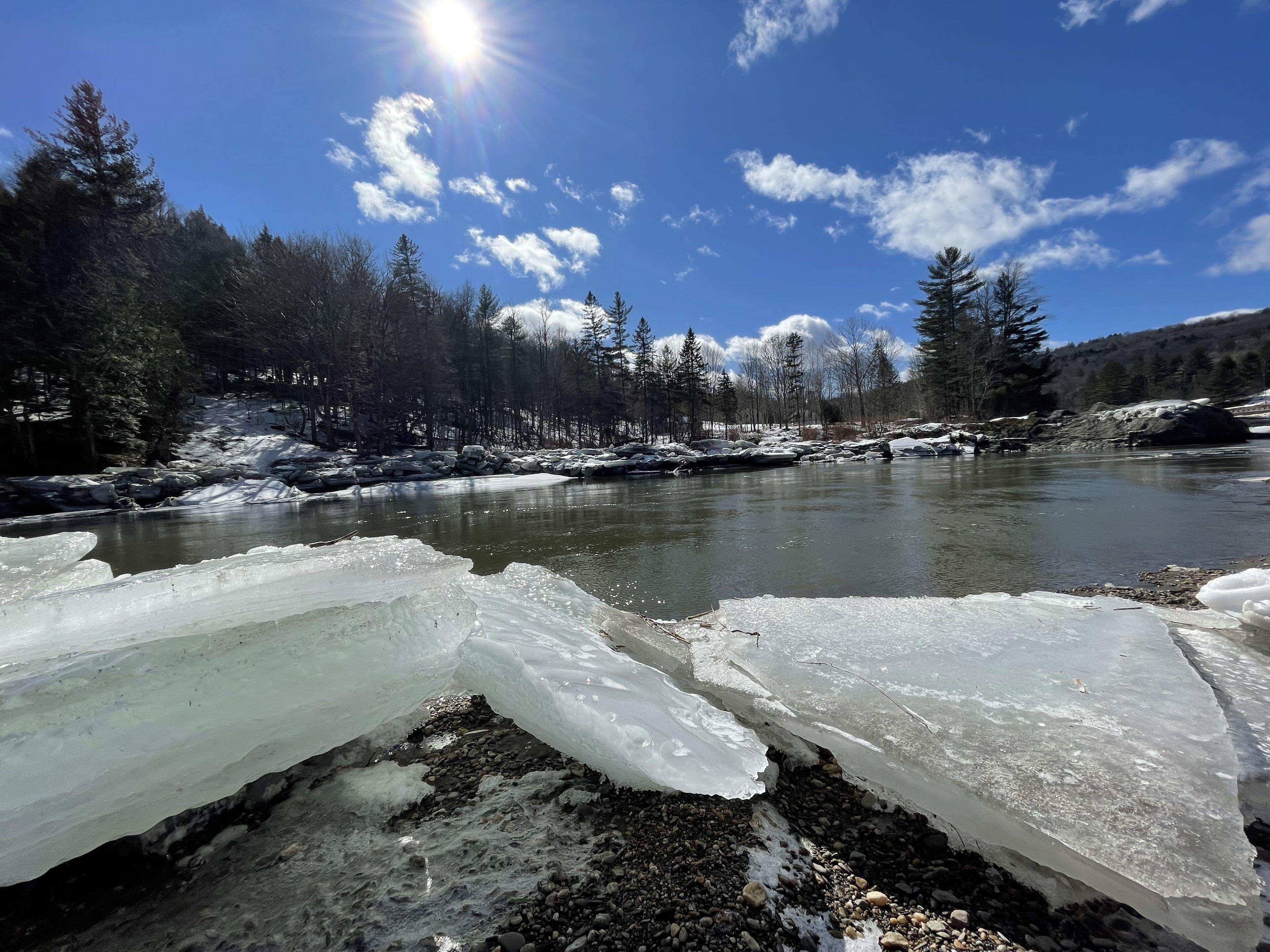 Ein Fluss mit Eisformationen und einer bewaldeten Landschaft unter blauem Himmel.