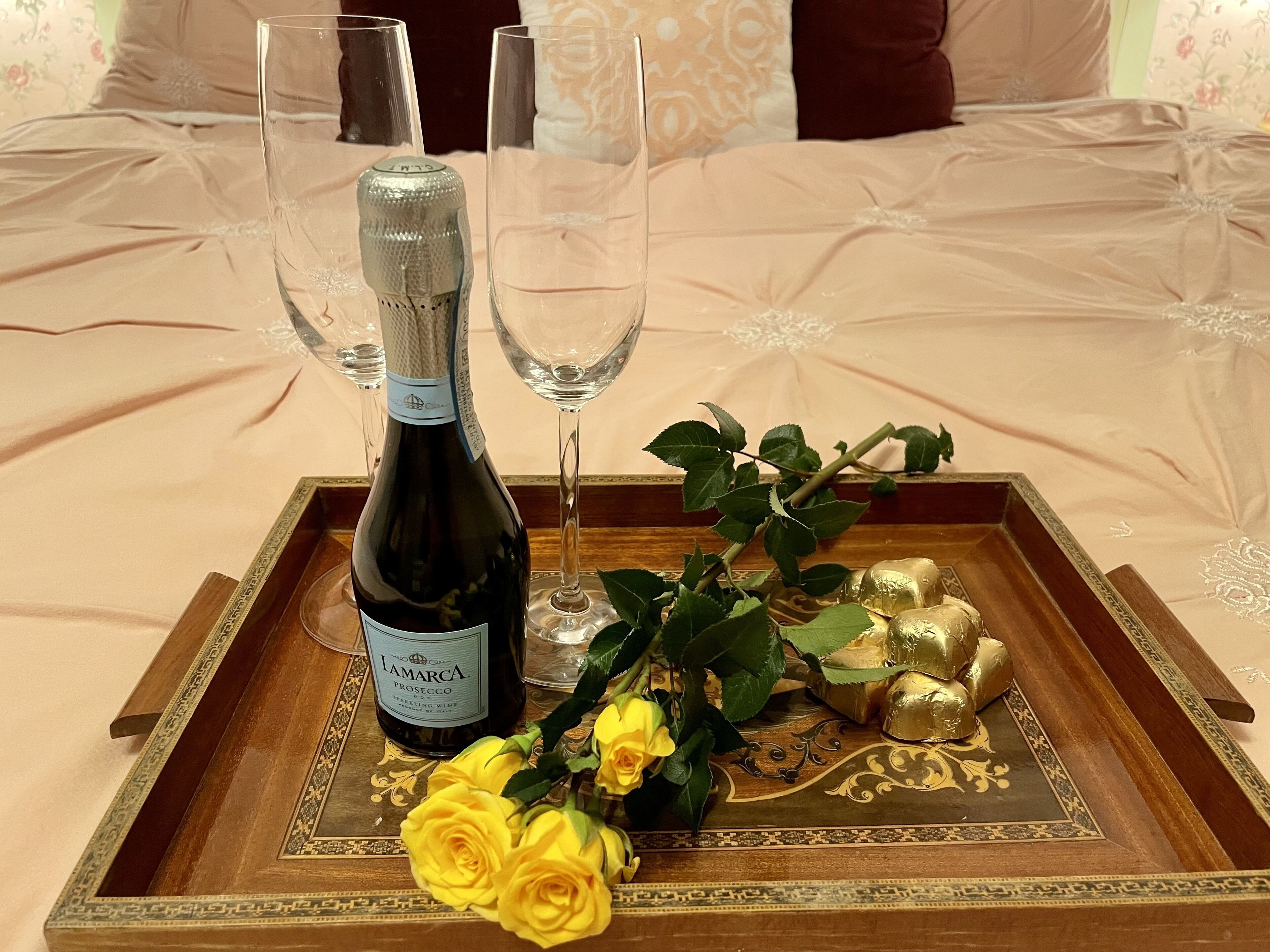 Una botella de vino espumoso Lamarca Prosecco, dos copas de vino vacías, un ramo de rosas amarillas y bombones de chocolate dorado sobre una bandeja de madera.