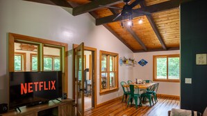 Una sala de estar acogedora con una televisión mostrando “NETFLIX”, pisos de madera, un ventilador de techo y un comedor con sillas verdes.