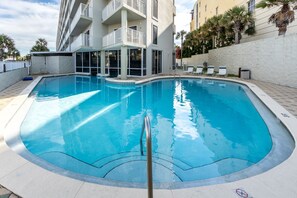 A swimming pool with a clear blue water, surrounded by a white building with balconies and palm trees.