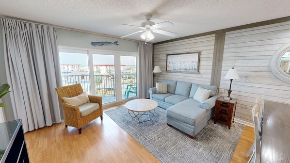 A spacious living room with a large window, a ceiling fan, a light-colored sofa, a wicker chair, and a small round table.