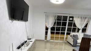 Ein modernes Wohnzimmer mit einem Flachbildfernseher, einem weißen Konsolentisch, einem Holzstuhl und einem großen Fenster mit weißen Vorhängen.