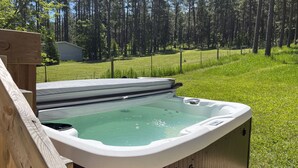 A hot tub with a wooden deck and a grassy yard with a fence and trees in the background.