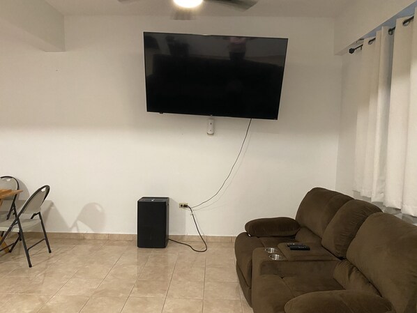 A living room with a flat-screen TV mounted on the wall, a brown sofa, a small table with a remote control, and a speaker on the floor. - El Yunque House with Private Pool & Amazing Views (Río Grande)