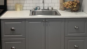 Una cocina con armarios grises, un salpicadero blanco de azulejos, un lavamanos de acero inoxidable y una pequeña planta decorativa.