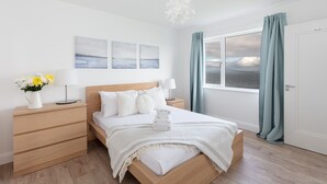 Un dormitorio moderno con cama de madera, ropa de cama blanca, una mesita de noche con lámpara, y vista al mar a través de una ventana con cortinas.